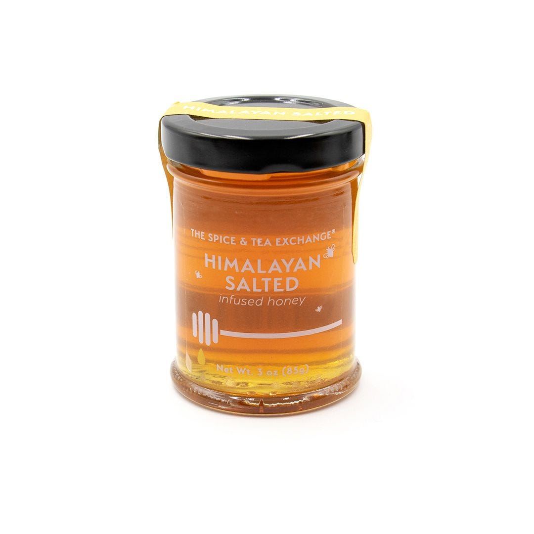 The Himalayan Salted Honey Jar is a 3 oz (85 g) glass jar with a black lid and yellow label showing a honey dipper. Honey drips down the side. Perfect for a cheese board. Background is white.